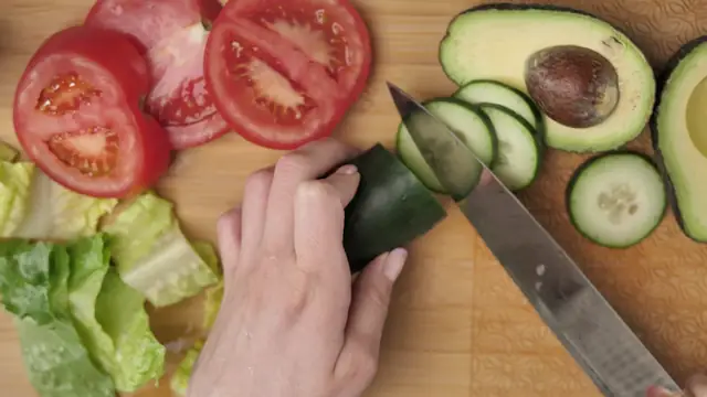 Hands slicing cucumber next to tomatoes, avocado and lettuce on a wooden board